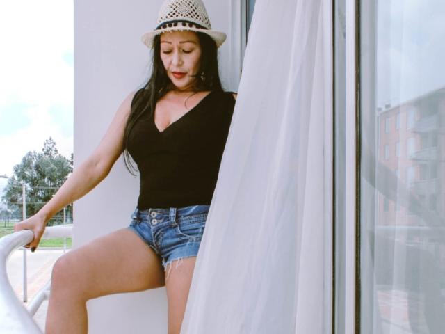 GinaOne with long black hair, wearing a black top, denim shorts, and a white straw hat, standing by a glass door with white curtains, showing her ass.