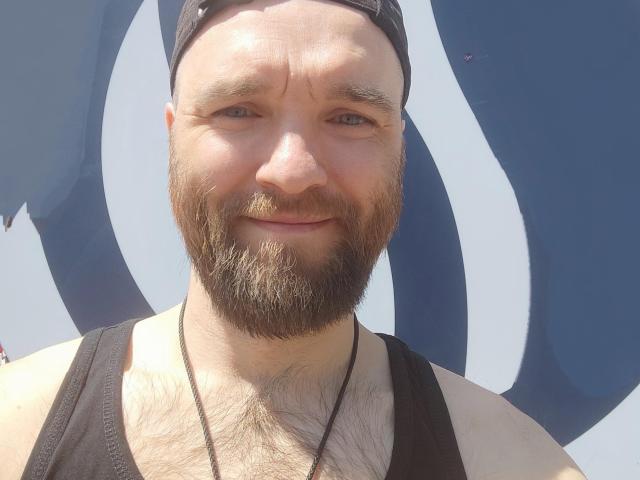SunColifild has a beard, black tank top, and black cap, with visible chest hair and a necklace, smiling in a sunny photo.