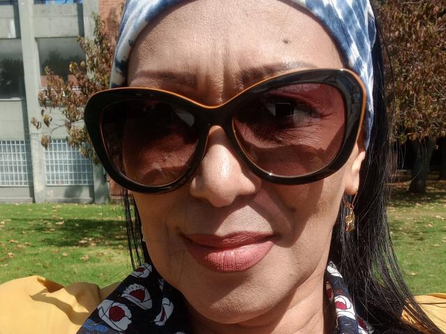 GinaOne's face, squinting eyes, large brown-tinted sunglasses, blue and white patterned headscarf, brown skin, yellow shirt with red and white pattern, and slightly parted pink lips.