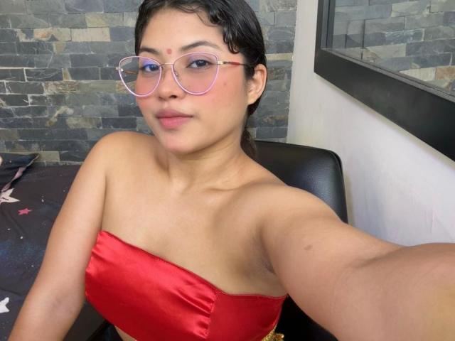 ShailaYojana with short curly black hair, wearing pink glasses and a red strapless top, showing small tits and a confident smile, sitting in a black chair.