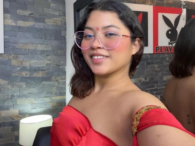 ShailaYojana with pink glasses, red off-shoulder top, and a cute smile, showing her tits in a selfie. She has a red dot on her forehead.