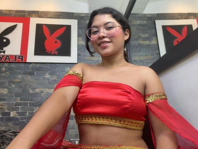 ShailaYojana with glasses, red satin top with gold trim, and red sheer sleeves, smiling, showing her small tits and dark hair, standing in front of Playboy art.