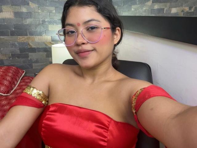 ShailaYojana with brown skin, red dress, gold arm bands, pink glasses, and a red dot on her forehead, smiling at the camera, showing her tits.