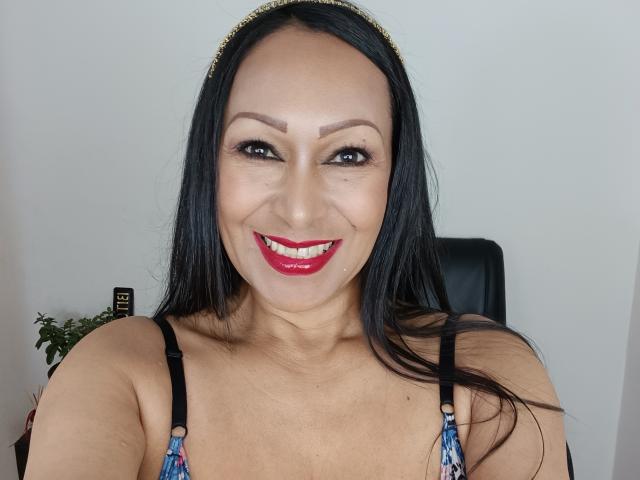 GinaOne has long black hair, brown eyes, red lipstick, and a gold headband. She's smiling with visible cleavage in a colorful, patterned top. Her tits are slightly visible.