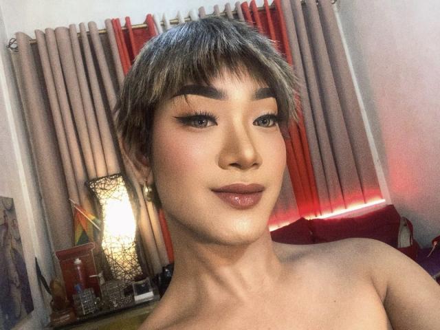 AsianHugeCockxX with short brown hair, thick eyebrows, winged eyeliner, and glossy pink lips, smiling with bare shoulders visible, standing in front of beige and red curtains.