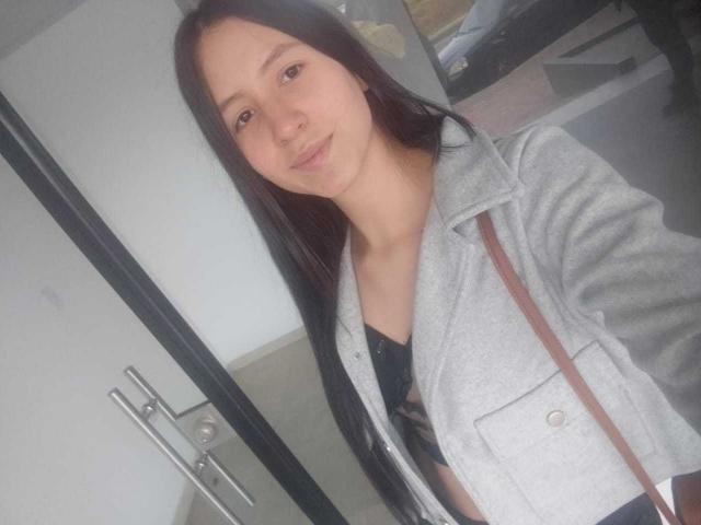 KarlaaGil met lang zwart haar, lichte huid, kleine borsten, gekleed in een grijze jas over een zwarte top, staand binnen, met een lichte glimlach, bruine band zichtbaar, selfie..