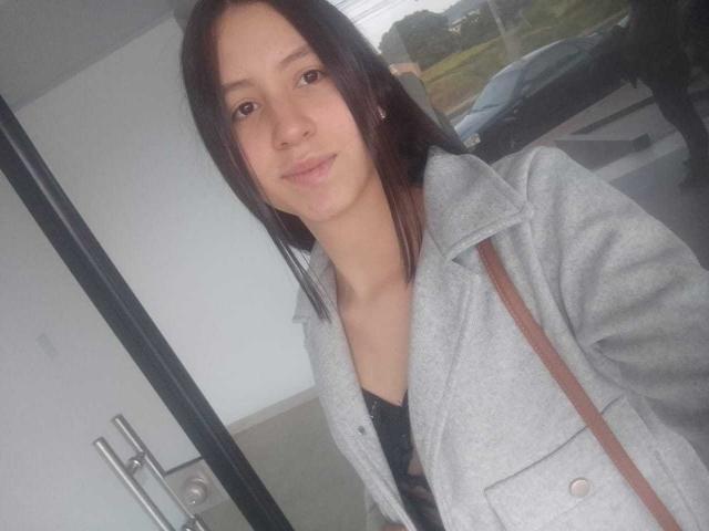 KarlaaGil, con cabello castaño largo, piel clara, pechos pequeños, vestida con un abrigo gris sobre una blusa negra, con una correa marrón, de pie en un interior, con una ligera sonrisa, mirando a la cámara..
