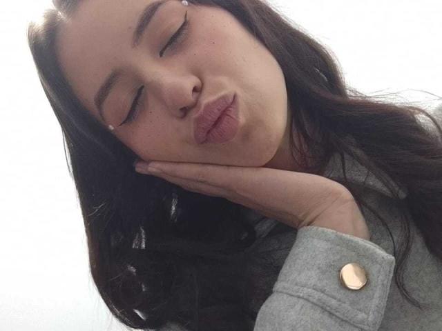 KarlaaGil with closed eyes, puckering her lips, holding her cheek with her hand, wearing a gray shirt with a gold button, has long dark brown hair.