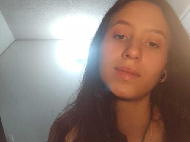 KarlaaGil hat langes, dunkelbraunes Haar, braune Augen und ein leicht gerötetes Gesicht mit glänzenden, rosafarbenen Lippen. Sie trägt kleine Ohrstecker und eine Halskette. Helles Licht von der Decke..