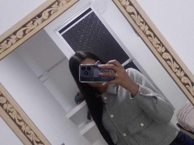 KarlaaGil tomando una foto de sí misma en el espejo, vistiendo una camisa gris abotonada, con su cabello negro cubriendo parte de su rostro, sosteniendo un teléfono con un accesorio pop socket, y mostrando ligeramente su trasero..