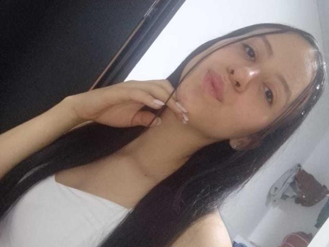 KarlaaGil, avec ses longs cheveux noirs, ses lèvres pulpeuses et son haut blanc sans bretelles, se touche le menton. Elle a la peau claire et fait une moue de baiser..