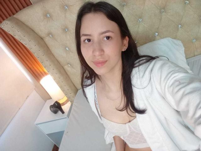 KarlaaGil met donker haar, lichte huid, kleine borsten in een witte kanten bh, een open witte badjas, zittend op het bed, kijkend naar de camera, een subtiele glimlach, een nachtlampje aan de linkerzijde..