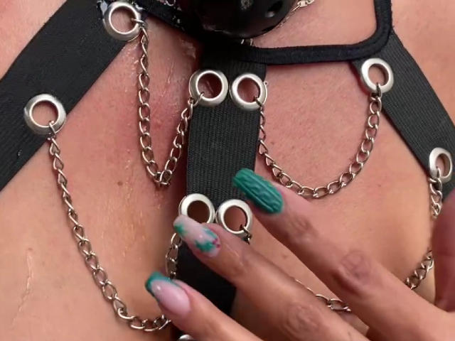 LailaCastillo's tits in black leather harness with silver chains, showing off her green and pink, textured nail polish, touching her chest.