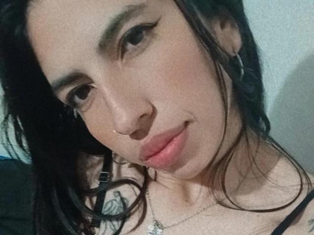 Andrushka's face with black hair, brown eyes, and a small nose ring. She's wearing a black top, a necklace, and has a tattoo on her right shoulder. Smiling subtly.