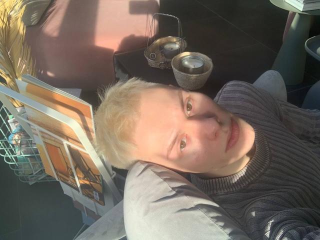 Matt Patrick mit kurzen, blonden Haaren, heller Haut und einem leichten Schnurrbart, trägt einen grauen, gerippten Pullover und liegt da. Sonnenlicht scheint auf sein Gesicht. Im Hintergrund sind Kerzen und Zeitschriften zu sehen..