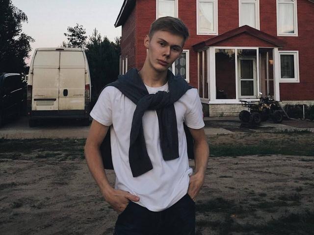MattPatrick stands in front of a red house, wearing a white t-shirt with a gray scarf, hands in pants pockets, looking serious, with a white van and bikes behind.