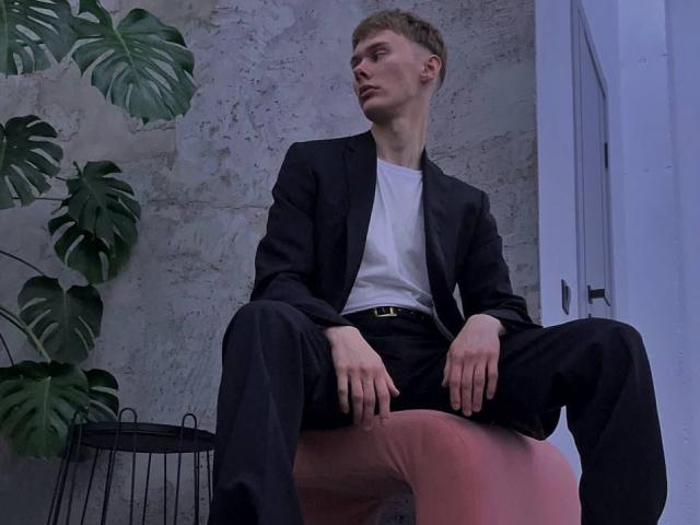 MattPatrick is sitting with legs spread, wearing a black suit over a white tee, and pink pants, looking to the side, with a plant and a wire basket nearby.