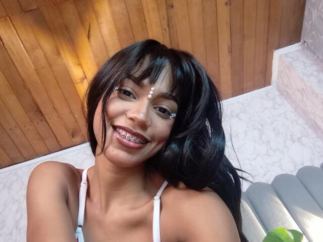 AylaCallowey with dark hair, white top, smiling, showing metal braces, white facial markings, wood and marble background, sitting on gray inflatable.