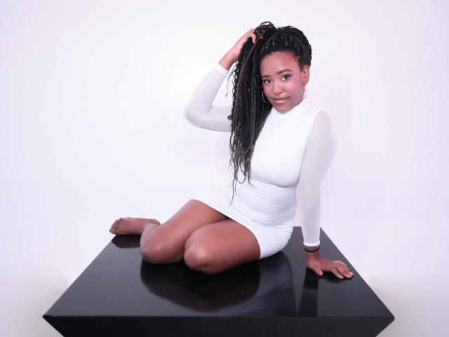 NaomiReyes, dark-skinned woman, sitting on black table, wearing white long-sleeve dress, holding braided black hair, smiling, bare feet, legs folded, reflective surface.