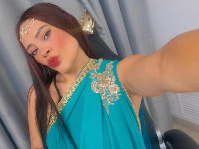 ShantalaKrisha with long brown hair, wearing a teal sari with gold embroidery, and a jeweled headpiece, pouting red lips, taking a selfie with a raised arm.