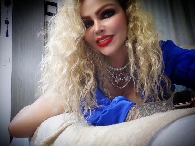 AnnaRichie, com cabelo loiro cacheado, batom vermelho, vestido azul, colar de pérolas, braço tatuado, segurando um telefone, sorrindo, apoiada em um travesseiro branco, sexy, atraente, mostrando seios grandes..