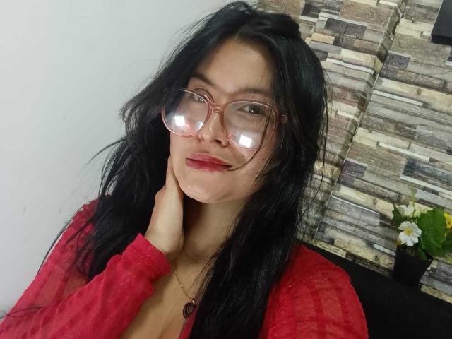 MeganHotCole with long black hair, red glasses, red lipstick, red sheer top, hand on neck, necklace, dark skin, and reflective glasses, showing off her cute face and tits.