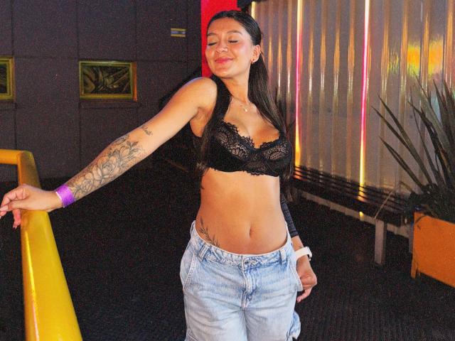 Thalia Ares, con un sujetador de encaje negro que deja ver sus pechos, un brazo tatuado y jeans claros, sonriendo con los ojos cerrados, apoyada en una barandilla amarilla. Lleva una pulsera morada..