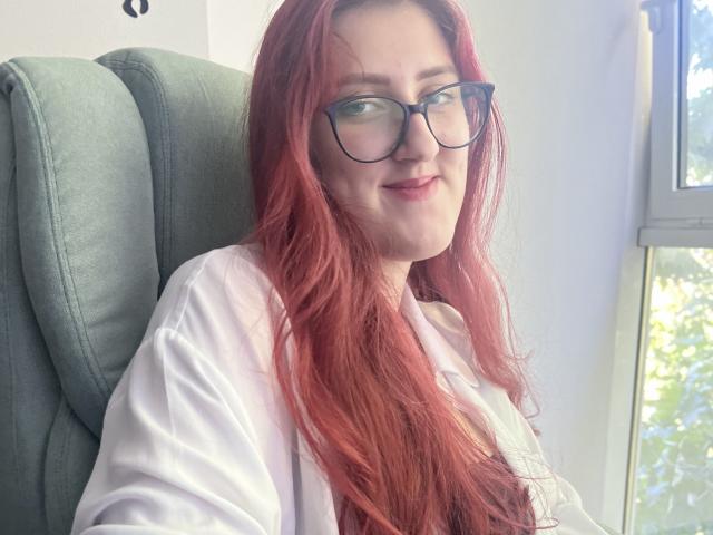 Triziya with red hair, glasses, and white shirt, sitting in a green chair, smiling slightly. Her tits are visible under the shirt. Sunlight from a window on the right.