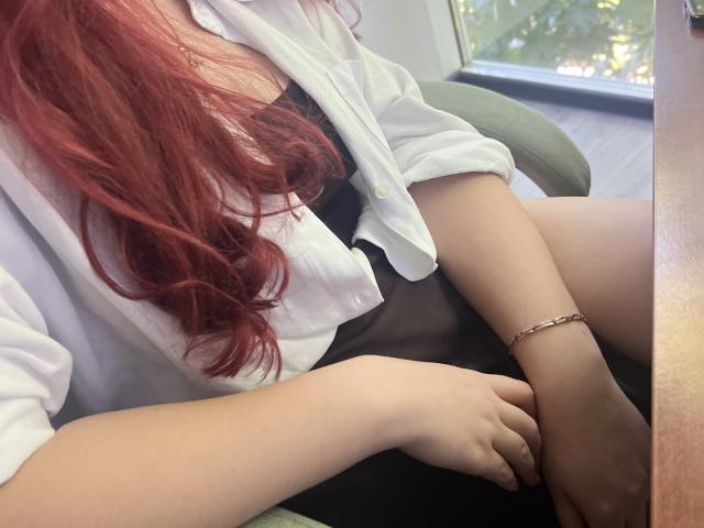 Triziya, com cabelo ruivo, camisa branca, vestido preto e pulseira, sentada com as pernas cruzadas, mostrando suas coxas e braços claros, em uma cadeira verde perto de uma janela..