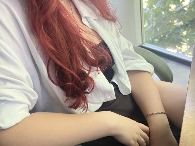Triziya, con cabello rojo, camisa blanca, vestido negro y pulsera, sentada junto a una ventana. Sus senos están cubiertos, su trasero es parcialmente visible. Su mano descansa sobre su muslo..