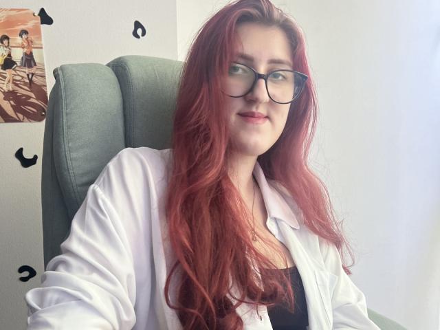 Triziya with red hair, glasses, white lab coat, black top, sitting in green chair, smiling, light skin, small ass, medium tits, slightly messy hair, casual, cute, confident.