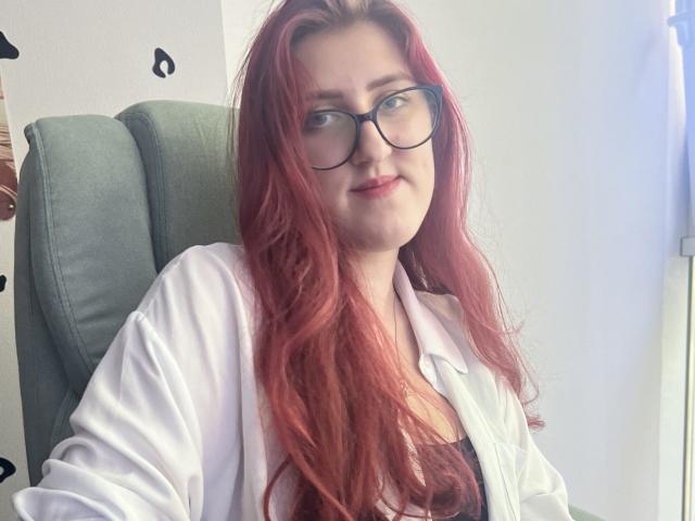 Triziya with long red hair, glasses, and a white lab coat, sitting in a green chair, smiling slightly, showing her tits through the coat.