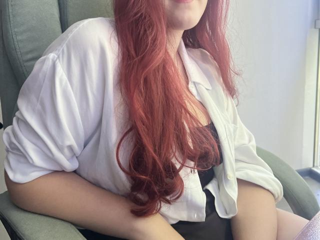 Triziya with red hair, wearing a loose white shirt, black top, and green chair, showing her tits and ass, sitting with arms on her thighs.