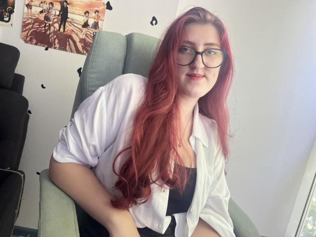 Triziya, con cabello rojo, gafas, camisa blanca y top negro, sentada con una ligera sonrisa, mostrando sus senos debajo de la camisa. Es linda y parece relajada..