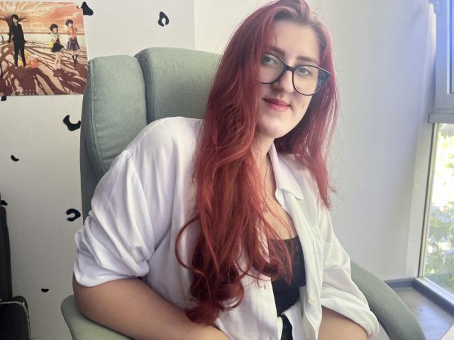 Triziya with long red hair, glasses, and a white shirt over black top, sitting in green chair, smiling, showing her tits slightly. Sunlight through window, relaxed.