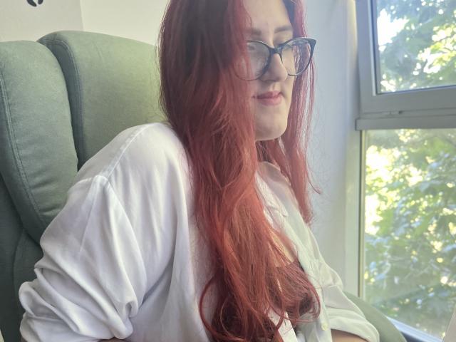 Triziya, com cabelo ruivo, óculos, camisa branca, sentada em uma cadeira verde, com luz do sol entrando pela janela, olhando para o lado, com a região do colo visível, expressão relaxada..