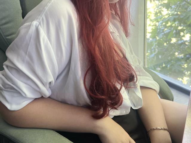 Triziya with long red hair, white loose shirt, green chair, and gold bracelet, sitting near window, showing her ass in black pants.