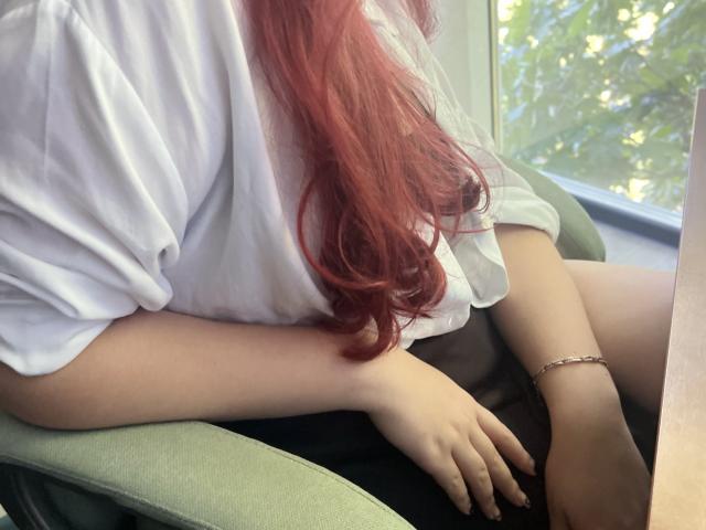 Triziya with red hair, wearing a white shirt, black shorts, sitting on a green chair, right arm resting, left arm on her knee, bracelet on her wrist.