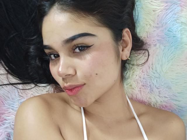 SaraLeonn with dark hair, brown eyes, and pink lips, wearing a white halter top, showing a small mole on her right cheek, lying on a fluffy, multicolored surface.