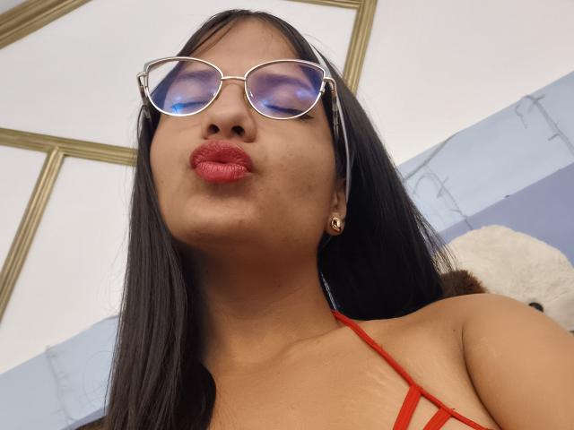 Laila Barker met rode lippenstift, volgehouden lippen, die een bril draagt, een rode top met bandjes, en gouden oorbellen, haar bruine haar is recht en lang, een kleine ringvormige oorbel is zichtbaar, haar ogen zijn gesloten..