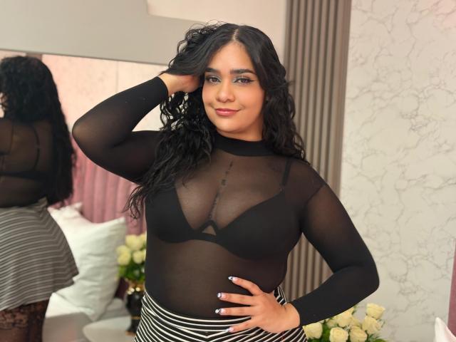 AliciaRelas with curly black hair, wearing a sheer black top revealing her tits and a striped skirt, standing with one hand in her hair, smiling confidently.