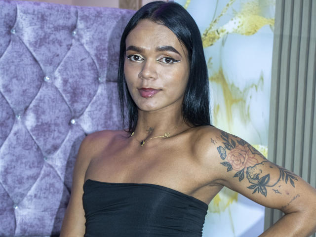 DulceDanesa has black strapless top, tattoos on right arm, straight black hair, brown eyes, subtle makeup, gold necklace, and purple tufted wall in background. She looks confident.