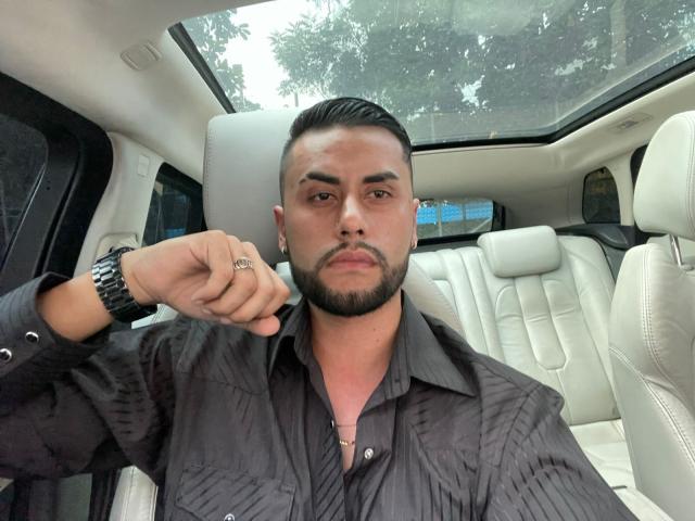 DammianConnors with a short, dark beard and slicked-back hair, wearing a black shirt, holding his fist to his chin, ring on finger, in a white car.
