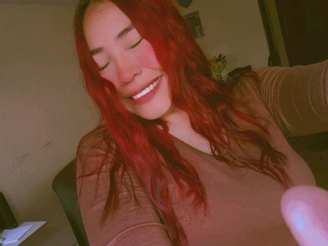 VenusSinclair with red hair, smiling, eyes closed, wearing a brown long-sleeve top, showing her tits slightly, in a selfie. Her hand is in the lower right corner.