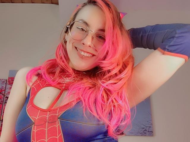 NatashaLau with pink hair, glasses, and Spider-Man costume, smiling, hand behind head, showing her tits in a blue and red suit, looking cute and sexy.