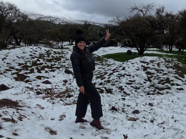 BelletinaXx stands in a snowy landscape, wearing a black jacket, pants, and boots with a beanie, making a peace sign with a smile, showing her ass slightly.