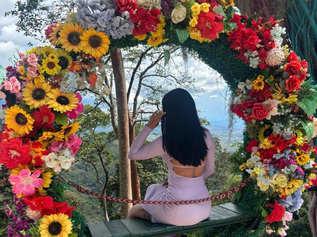 AnyaJay with long black hair, sitting on green bench, back to camera, in pink dress, looking through sunflower and rose floral arch, surrounded by colorful flowers, outdoors.