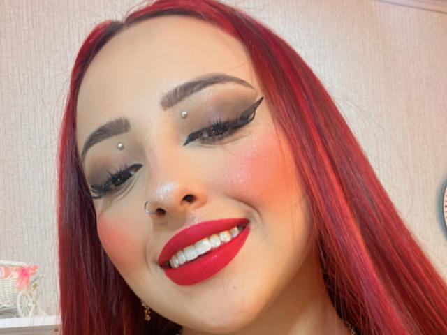 KylieCharm mit roten Haaren, auffälligem schwarzen Eyeliner und rotem Lippenstift, lächelnd mit gestochenen Augenbrauen, Nase und Oberlippe, wobei ihre roten Lippen und ihre glatte Haut zur Geltung kommen..