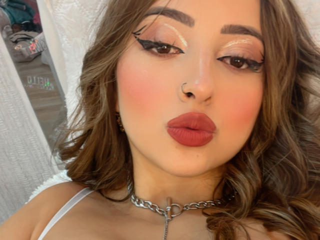 KylieCharm has brown wavy hair, a nose ring, and wears a metal chain collar with red lipstick and glossy eyes. Her makeup is bold with dramatic eyeliner.