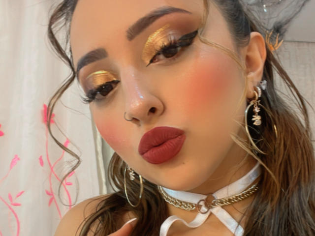 KylieCharm heeft goudkleurig oogmake-up, rode lippenstift, een witte choker en oorringen. Haar bruine haar is golvend, en ze frumelt met haar glanzende, roze lippen..